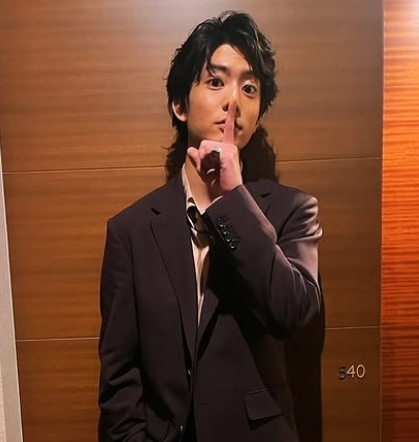 ブラックのスーツ姿で口の前で人差し指を立てて口を紡ぐようなしぐさで写っている伊藤健太郎