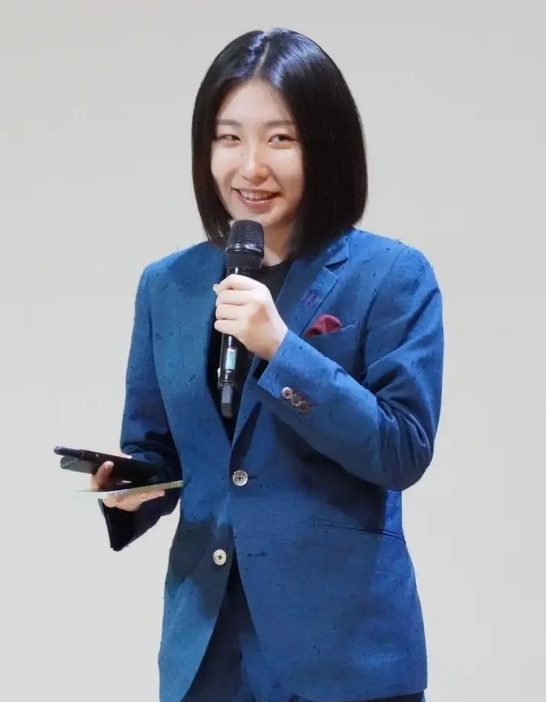 ブルーのジャケット姿でマイクを握り講演をしている姿の合田瞳