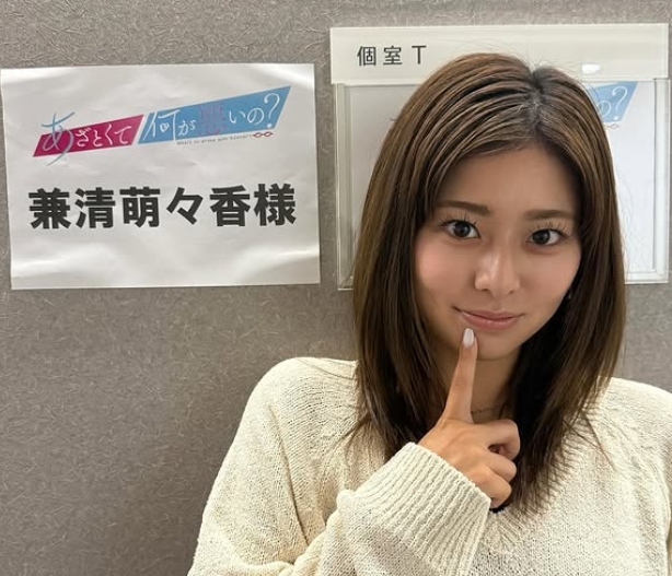 兼清萌々香さんがテレビ番組『あざとくて何が悪いの？』の控室前で、指を口元に添えてポーズをとっている様子。ライトカラーのニットを着用し、番組名と名前が書かれた案内板の前に立つ姿が印象的。