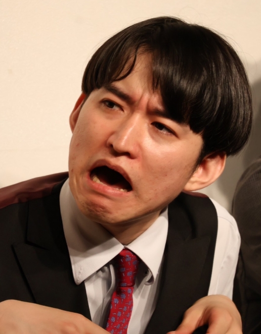 めぞんの吉野おいなり君が白シャツと黒ベスト、赤いネクタイ姿で口を半開きにした表情豊かにポーズをとっている写真