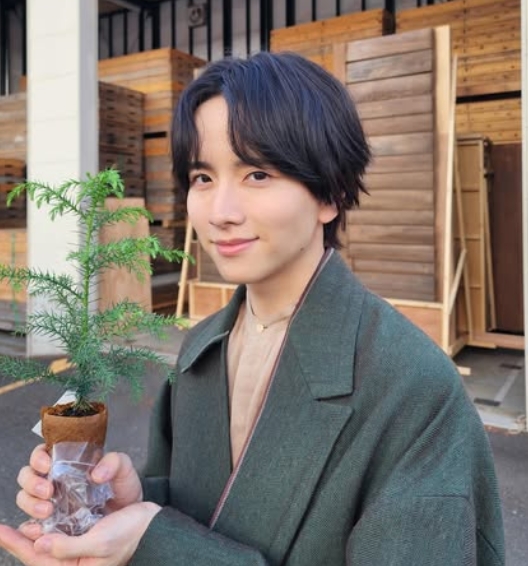 緑のコートを着た板垣李光人が小さな苗木を手に持ち、木材の背景の前で環境活動に参加している様子